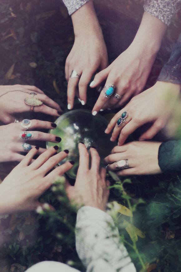 Many hands reaching inward to touch a crystal ball, all with different nail polish, rings, and sleeves visible.