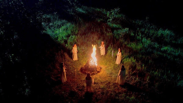 A group of people dressed in white, standing in a circle around a fire at night with lush greenery around them
