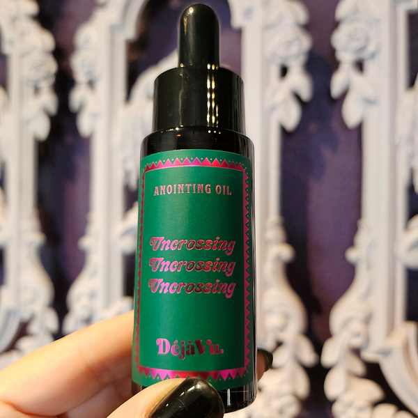 A hand holding a small black dropper bottle of anointing oil with a green label that reads 'Anointing Oil' and 'Uncrossing' in pink lettering, set against a purple and white ornate background.