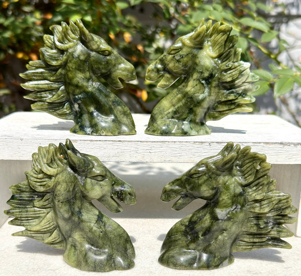 Four green stone horse head carvings displayed outdoors on a white surface, with leafy greenery and a light wall in the background.