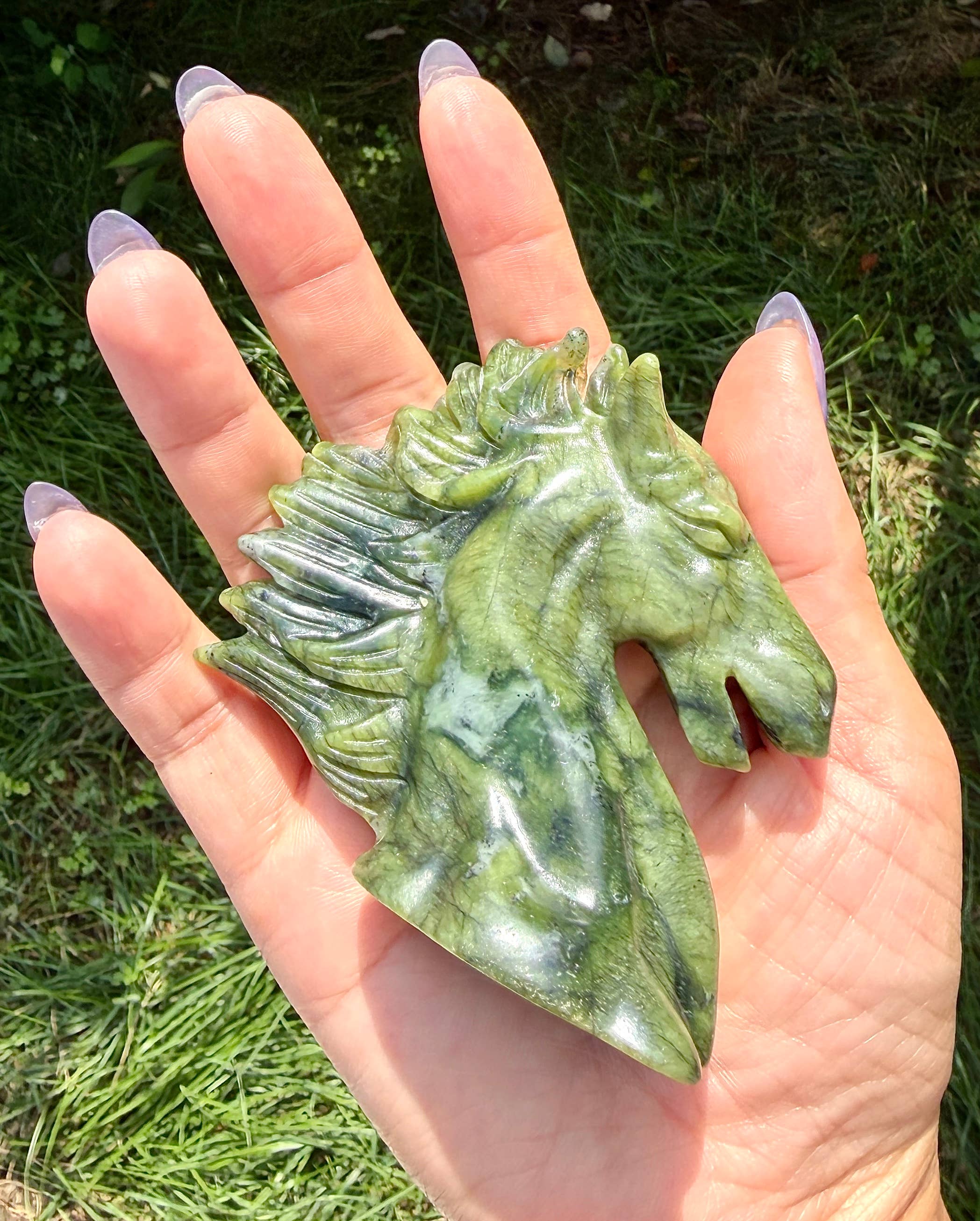 A hand with purple nail polish holds a polished green jade horse head carving outdoors against a background of green grass.