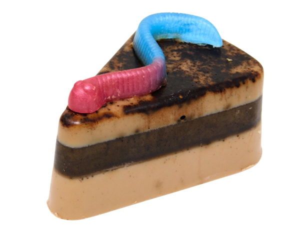 Graveyard Dirt Cake Soap - triangular slice-shaped soap with layered chocolate and caramel tones, real cocoa topping, and a pink and blue gummy worm embed on top, on a white background
