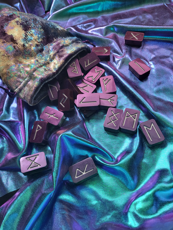 A set of purple rune tiles with silver engraved symbols scattered from a matching purple fabric pouch onto a shiny iridescent teal and purple draped background.