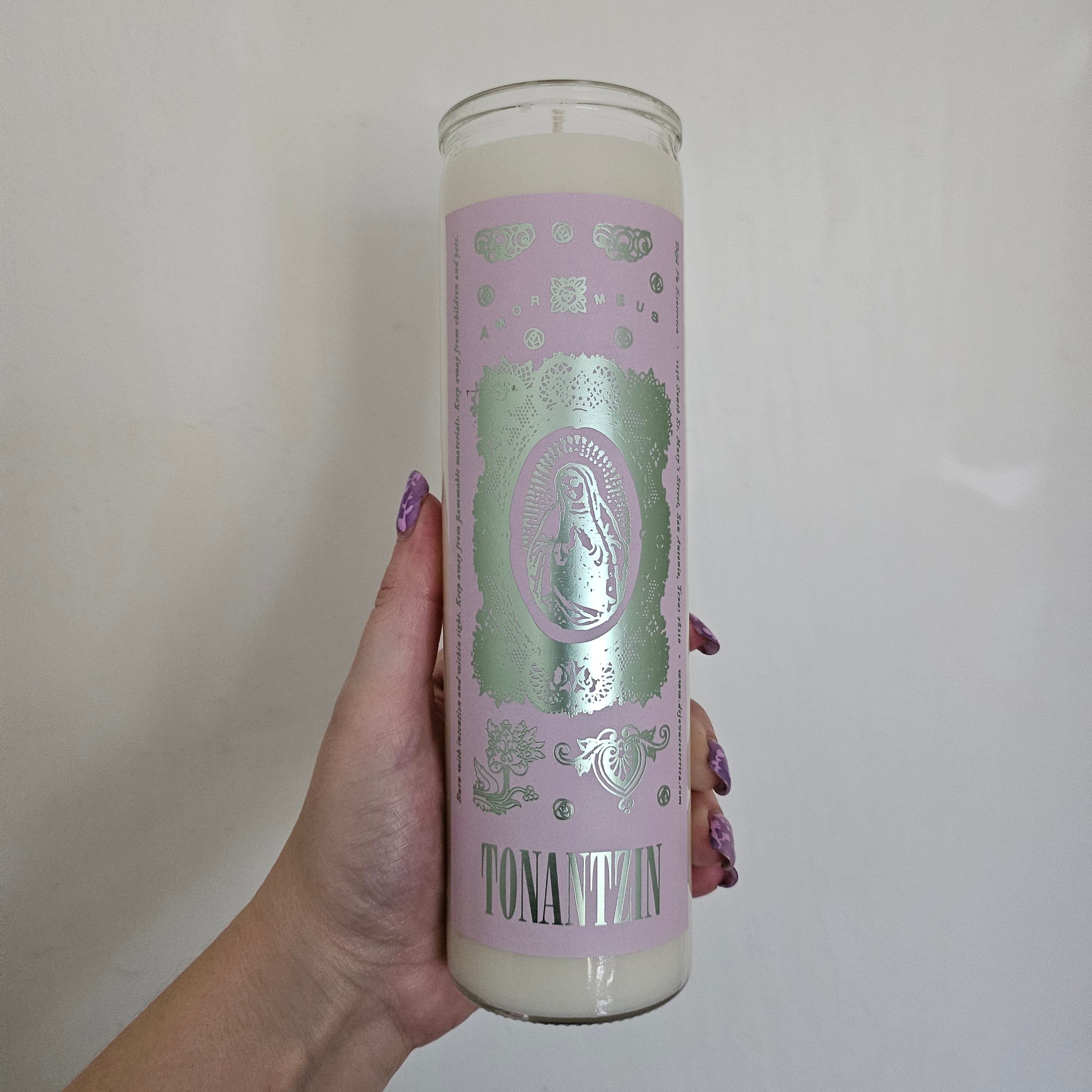A hand holding a white Tonantzin candle with a lavender label with mint green foil inside a glass container