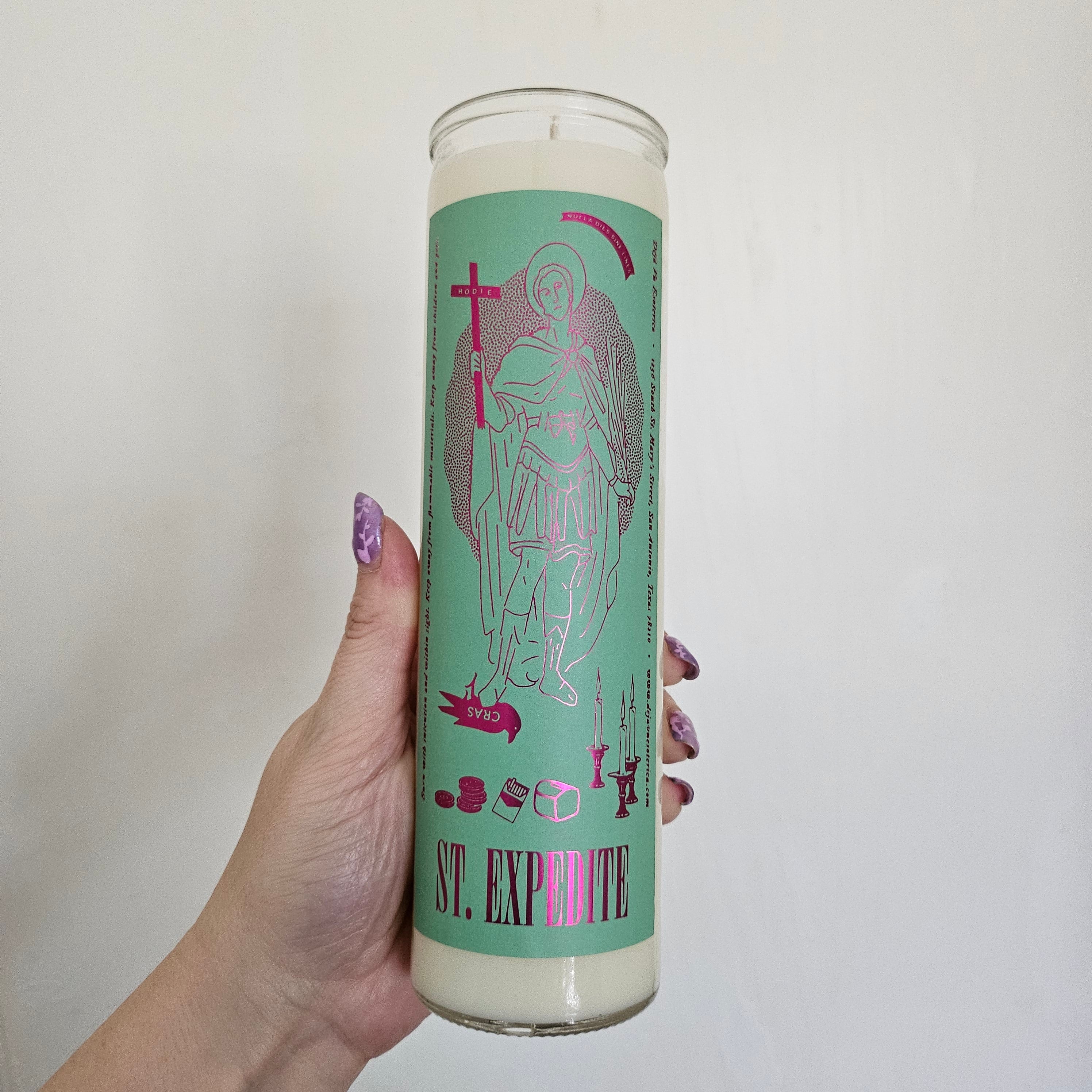 A hand holding a white St Expedite candle with a green label with magenta foil inside a glass container