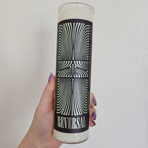 The label on this candle features multidirectional arrows in mint green foil stamped on a dark purple background, the word REVERSAL is at the bottom. A hand holds the candle, with white wax.