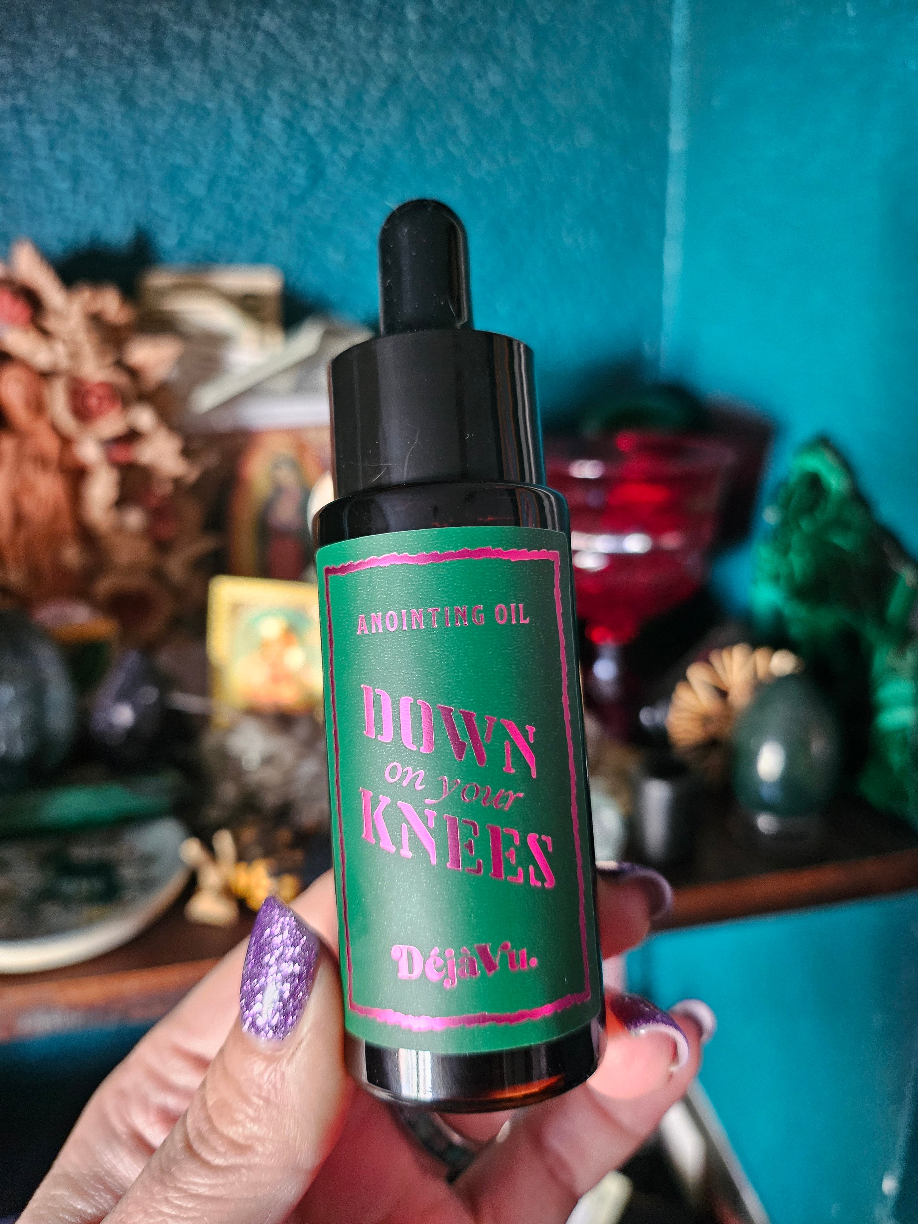 A hand with purple glitter nail polish holds a small amber dropper bottle labeled 'Down on Your Knees' by the brand Deja Vu. The background is a teal wall with blurred objects on an altar.