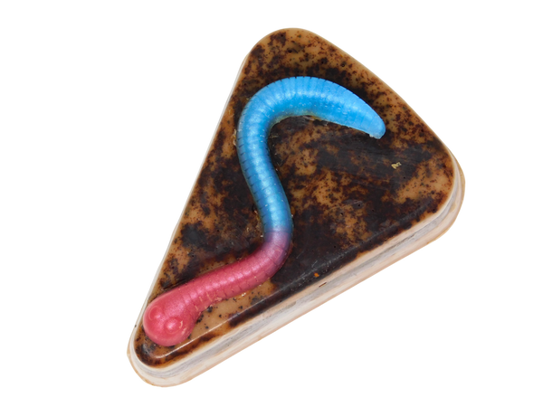 Graveyard Dirt Cake Soap - triangular slice-shaped soap with layered chocolate and caramel tones, real cocoa topping, and a pink and blue gummy worm embed on top, on a white background