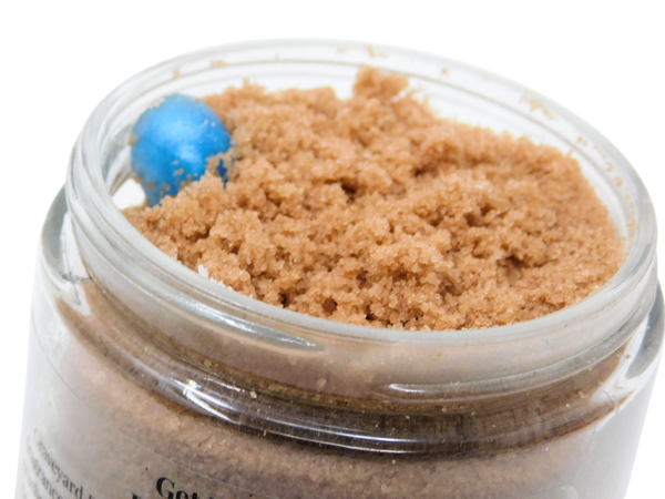 Close-up top-down view of Graveyard Dirt Cake Body Scrub showing the coarse cocoa sea salt texture and a blue soapy gummy worm in an open glass jar