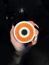 Somebody's Watching Me Evil Eye Soap - round soap with concentric orange and white rings and a black center pupil on a white background, held by a hand in a dark bath