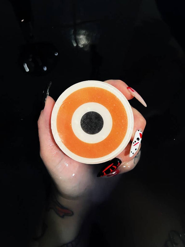 Somebody's Watching Me Evil Eye Soap - round soap with concentric orange and white rings and a black center pupil on a white background, held by a hand in a dark bath