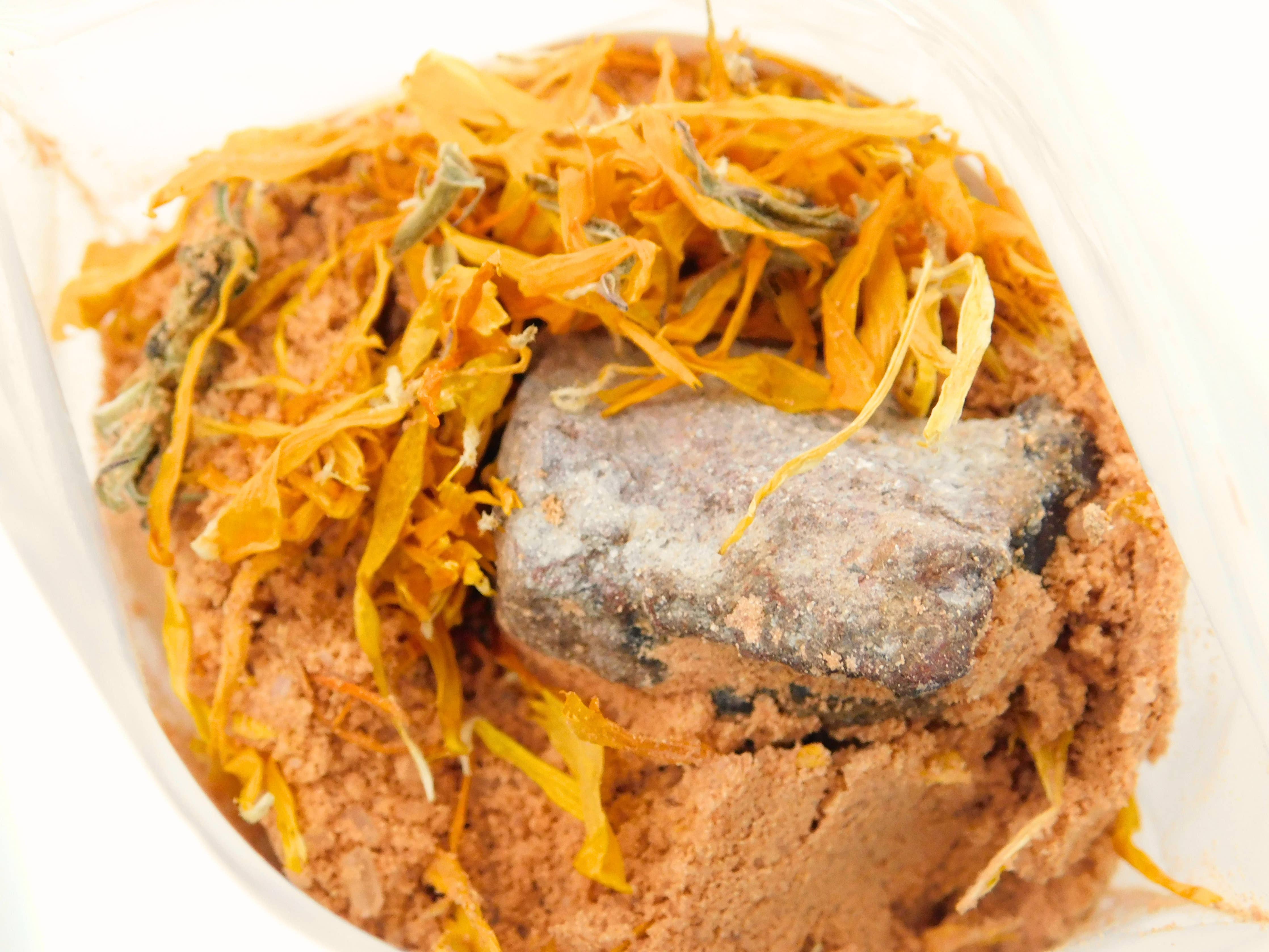 Close-up of Pumpkin Spell Bath Potion contents showing a raw fire agate chunk nestled in warm orange pumpkin clay powder topped with bright dried calendula marigold petals