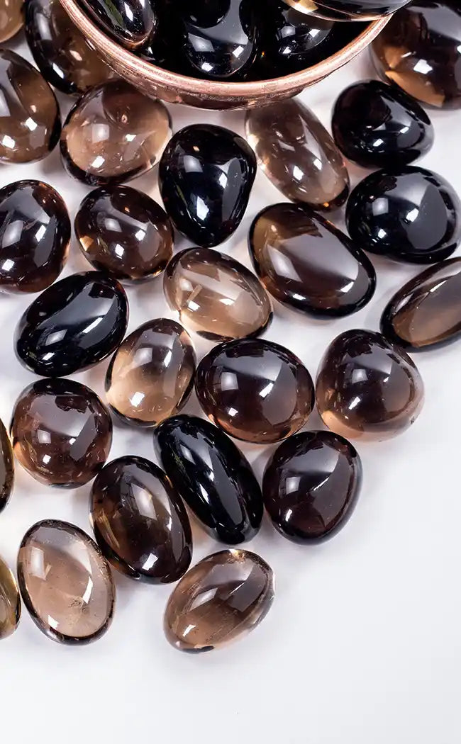 Tumbled smoky quartz gemstone tumbles in shades of brown, black, and translucent smoky gray, with some resting in a copper bowl on a white background.