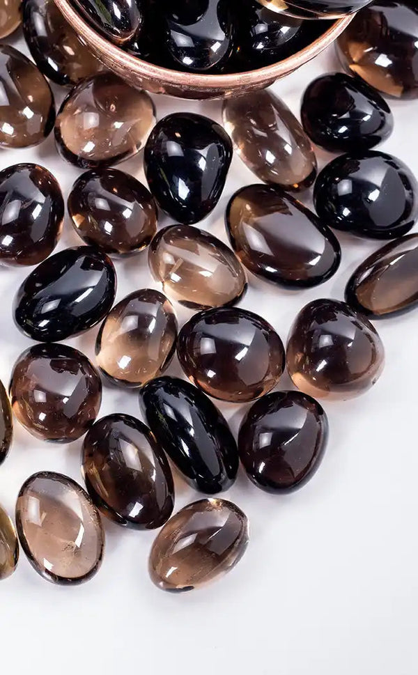 Tumbled smoky quartz gemstone tumbles in shades of brown, black, and translucent smoky gray, with some resting in a copper bowl on a white background.