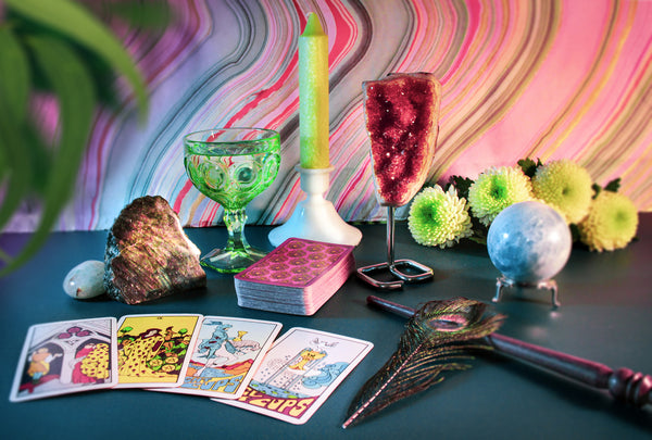 Tarot cards, crystals, flowers, a candle, magic wand, peacock feather, and glass chalice are carefully arranged against a marbled background and washed in soft light
