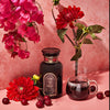 Ruby Moon tea in a dark glass apothecary jar sitting beside a steaming glass mug of deep red tea, sitting beside fresh cherries, red dahlias, and bougainvillea on a pink background
