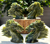 Four green stone horse head carvings arranged around a wooden pedestal, with a blurred outdoor background of foliage and light.