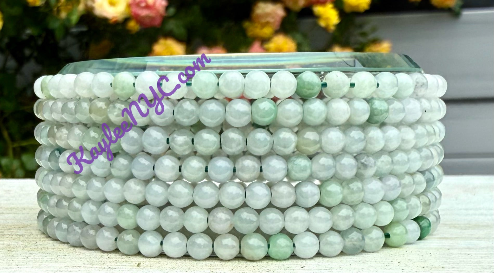 Stack of green beaded bracelets on a white surface with blurred flowers in the background