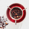 Loose leaf tea blend steeping in a silver infuser over a deep red glass cup, top-down view on white background