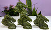 Three green stone horse head sculptures displayed on a white surface with a purple wall and potted plants in the background.