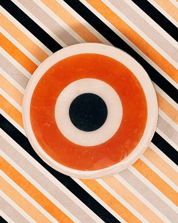 Somebody's Watching Me Evil Eye Soap on a diagonal orange, black, and cream Halloween stripe background, showing the full round orange evil eye design