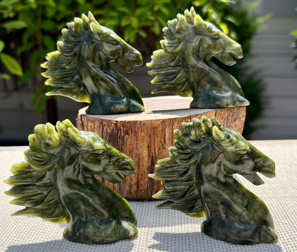 Four green stone horse head carvings arranged on and around a wooden block, photographed outdoors with blurred greenery and a light wall in the background.