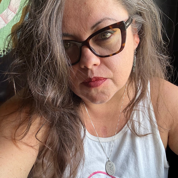 A close-up portrait of Elle, a woman with long grayish-brown hair wearing tortoiseshell cat-eye eyeglasses, a white tank top, and a silver pendant necklace.